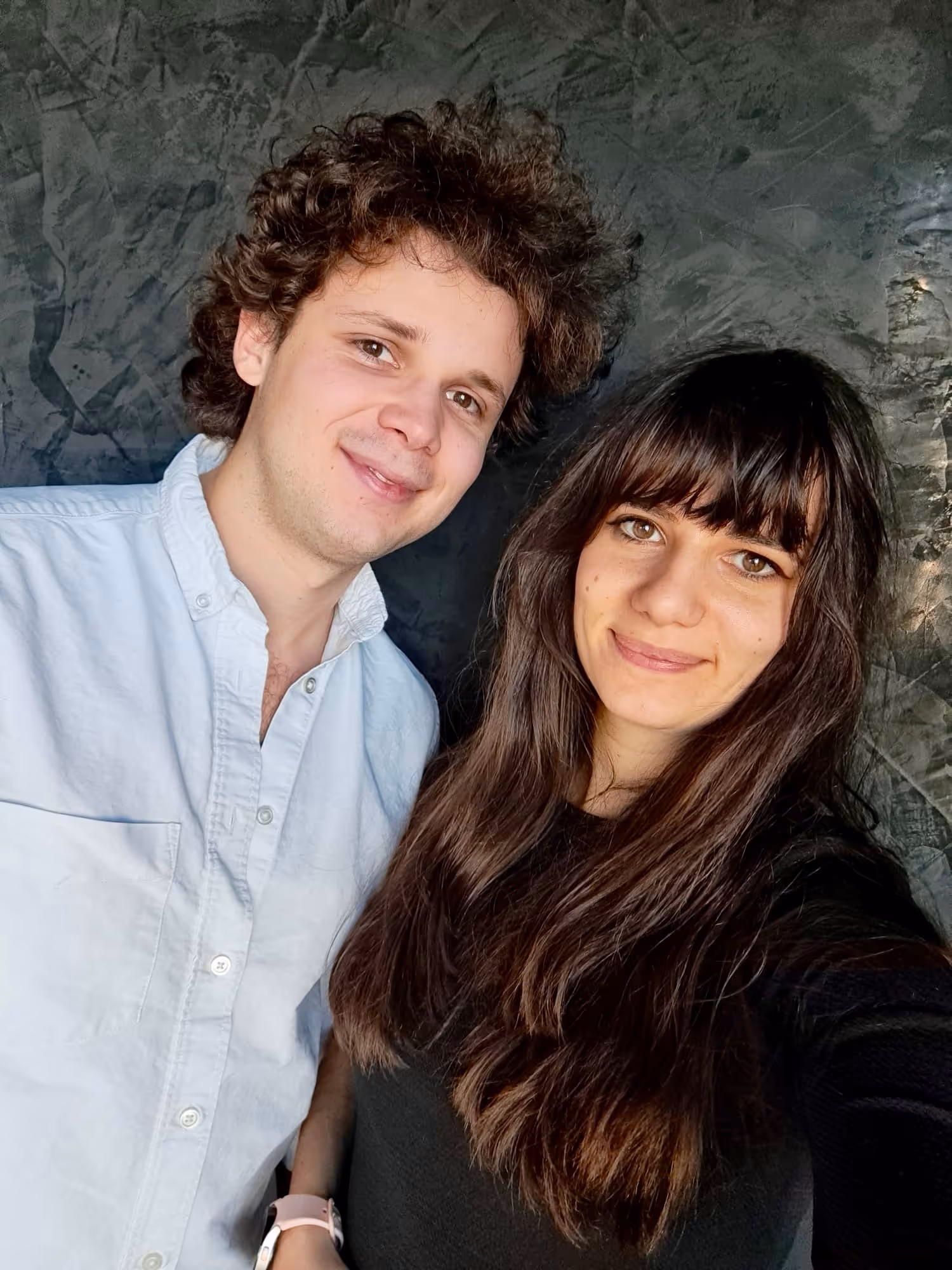 Young man and woman smiling together in front of a textured gray wall, close-up portrait.