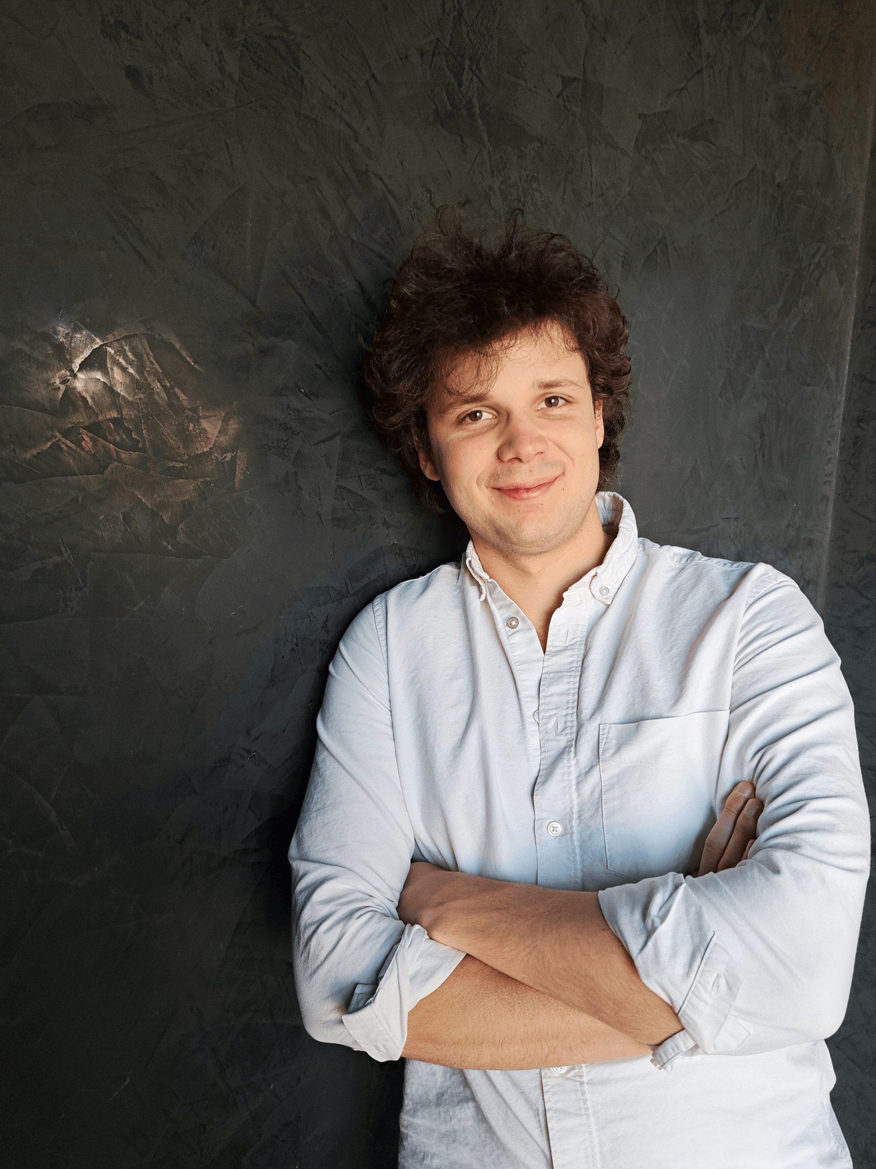 Young man with curly hair wearing a white button-up shirt, standing with arms crossed against a textured dark wall.