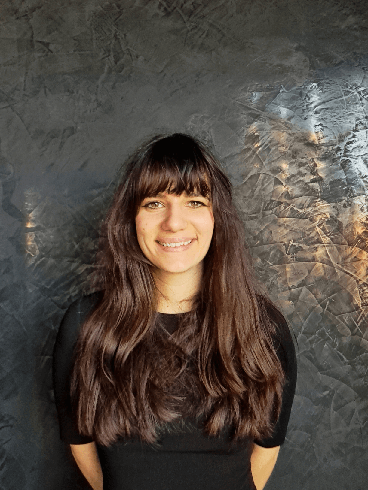 Smiling woman with long brown hair and bangs wearing a black top against a textured dark wall.