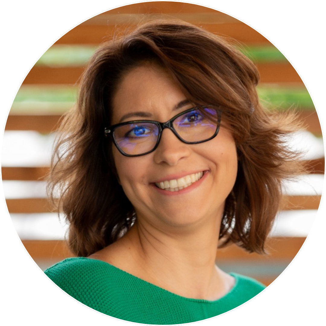 Smiling woman with shoulder-length brown hair wearing black glasses and a green top against a blurred wooden background.
