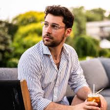 Man with dark hair and glasses wearing a striped shirt, sitting outdoors holding a drink with a straw.